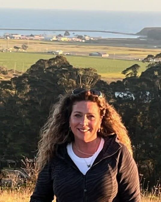 Vanessa Evans smiling in golden evening light on a foothill ridge, with a harbor and the Pacific coastline behind her